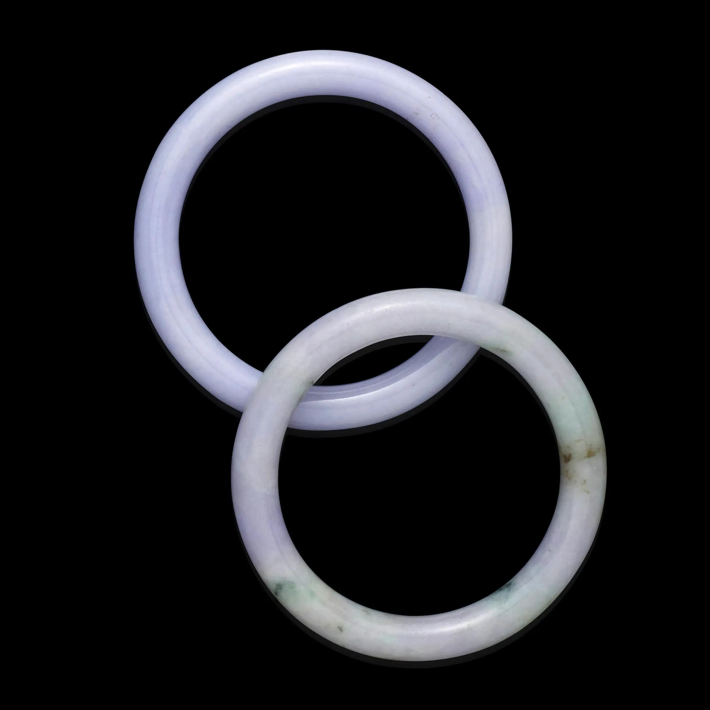 Pair of polished lavender jadeite bangle bracelets on black background; one evenly lilac, one lilac with subtle mottling